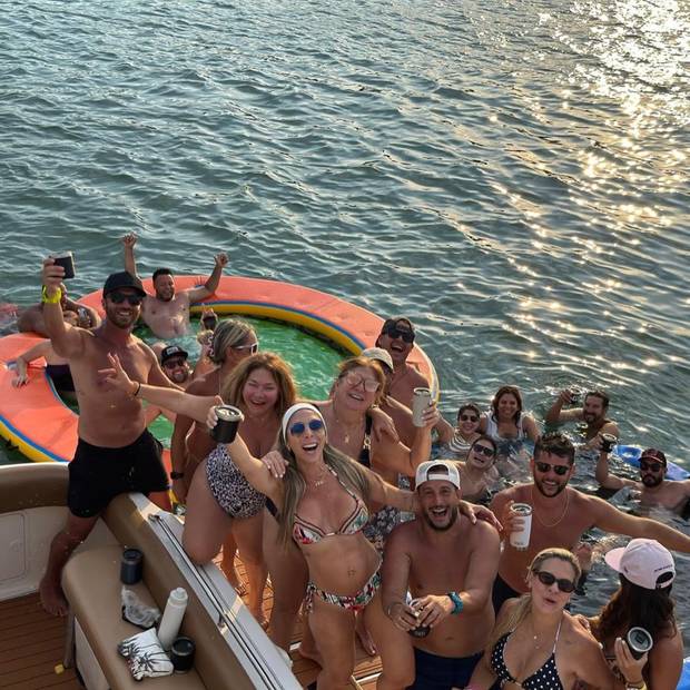 Un grupo grande en la popa con bebidas, y detrás una piscina flotante naranja sobre el agua iluminada